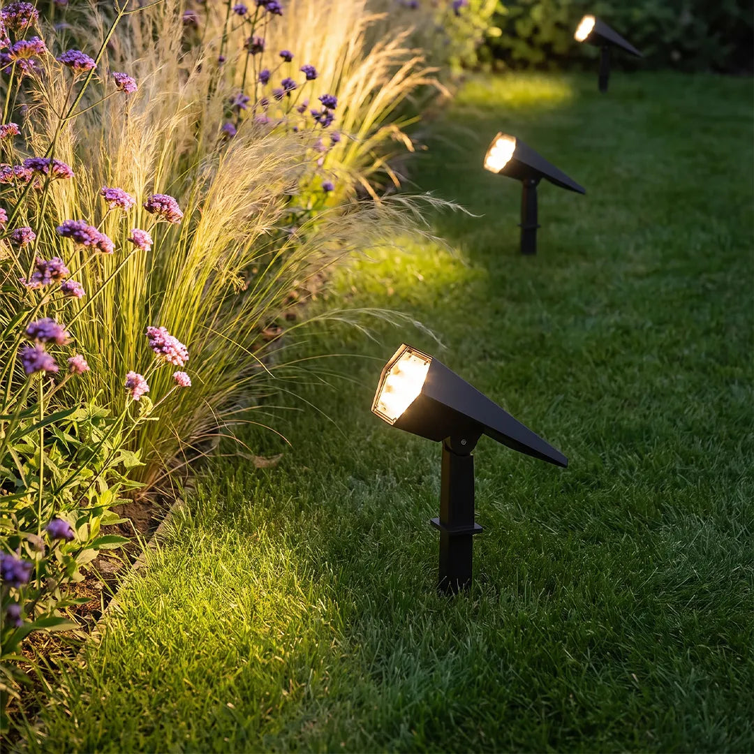 Flood spike light positioned beside flowering plants, providing focused landscape lighting along a grassy garden border.