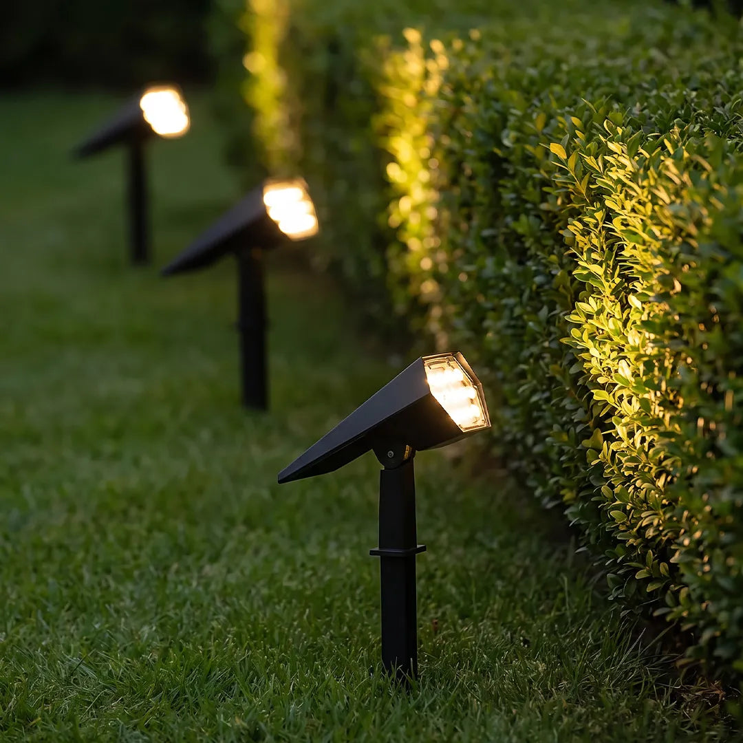 Flood spike lights installed along a garden hedge, casting warm illumination across the lawn and shrubs at night.
