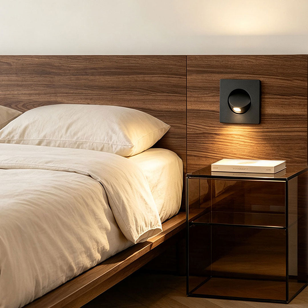 Modern flush mount wall light installed beside bed with warm ambient glow and wood panel backdrop.