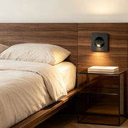 Modern flush mount wall light installed beside bed with warm ambient glow and wood panel backdrop.
