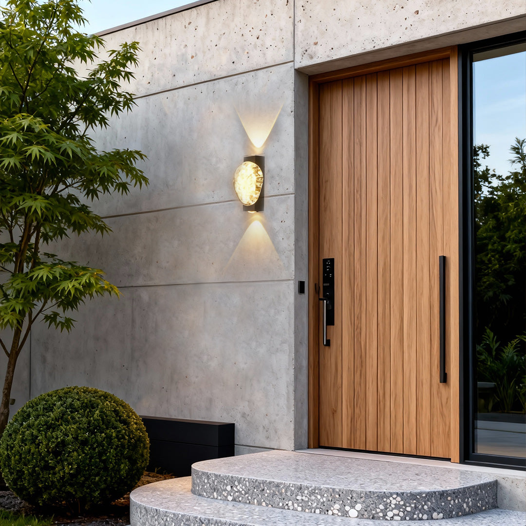 Gold up and down outdoor wall lights installed beside a wooden front door, casting warm accent lighting on a white wall.