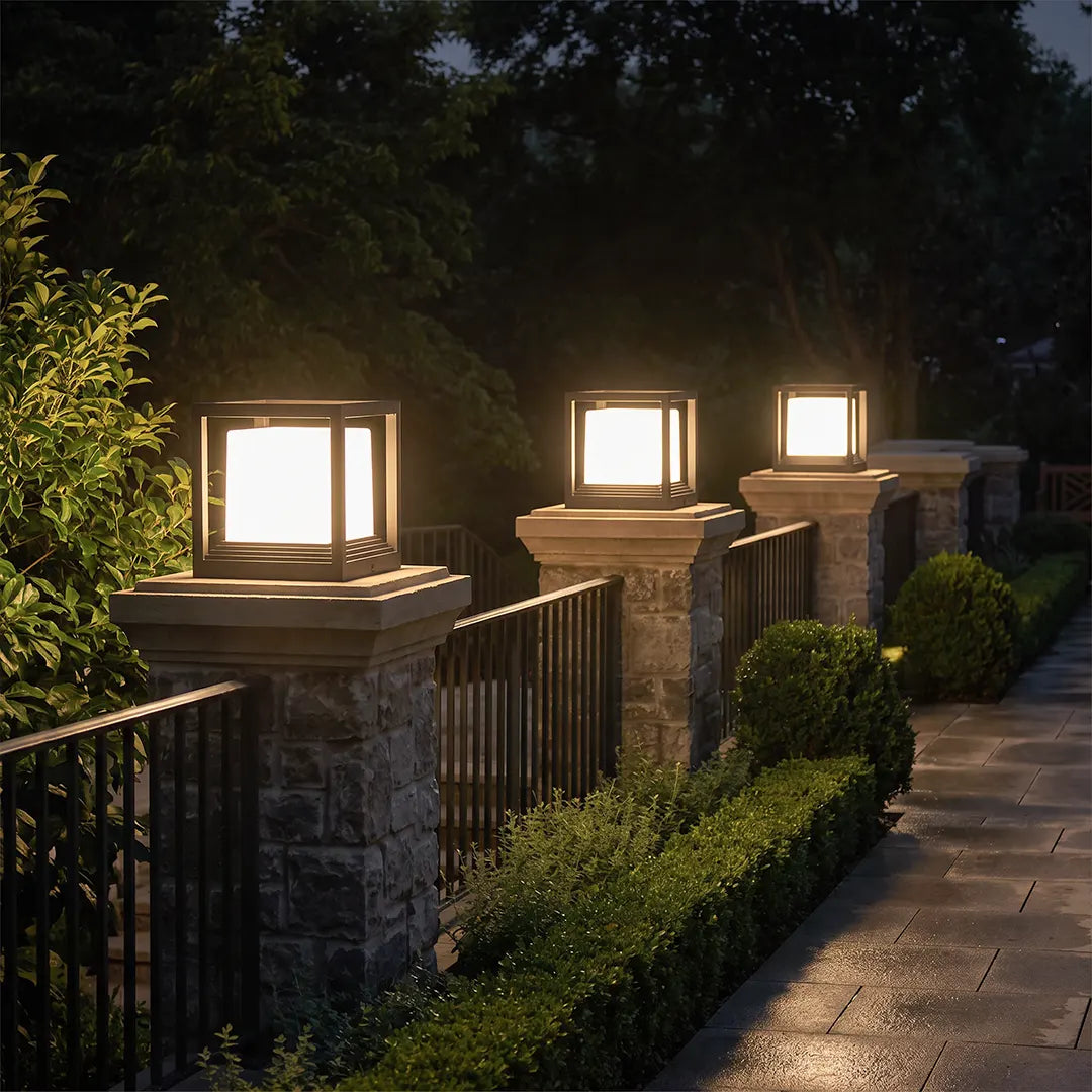 Garden pedestal lights mounted on stone columns along a landscaped walkway at night.