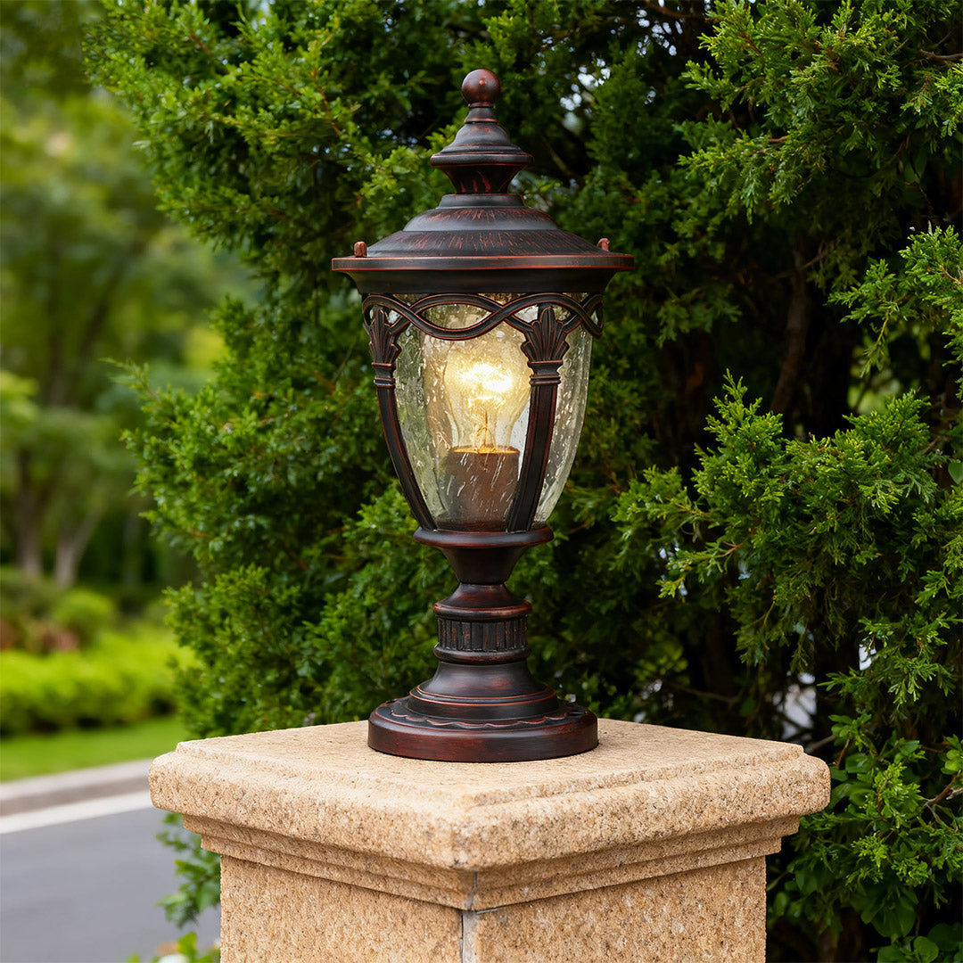 Gate pillar light standing on textured beige pedestal beside roadway with lush green hedge backdrop