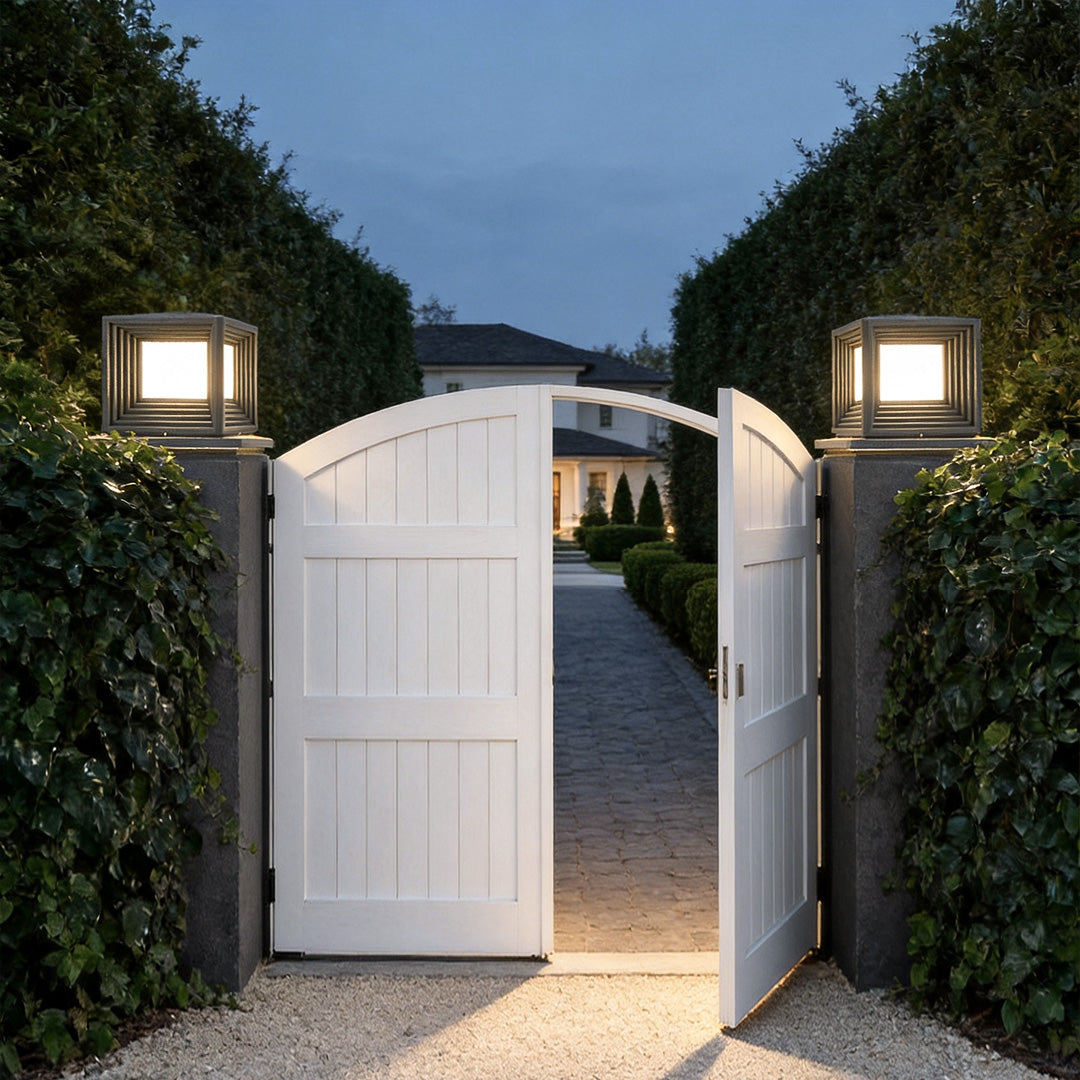 Modern gate pillar lights illuminating a white gate entrance with lush greenery at dusk.