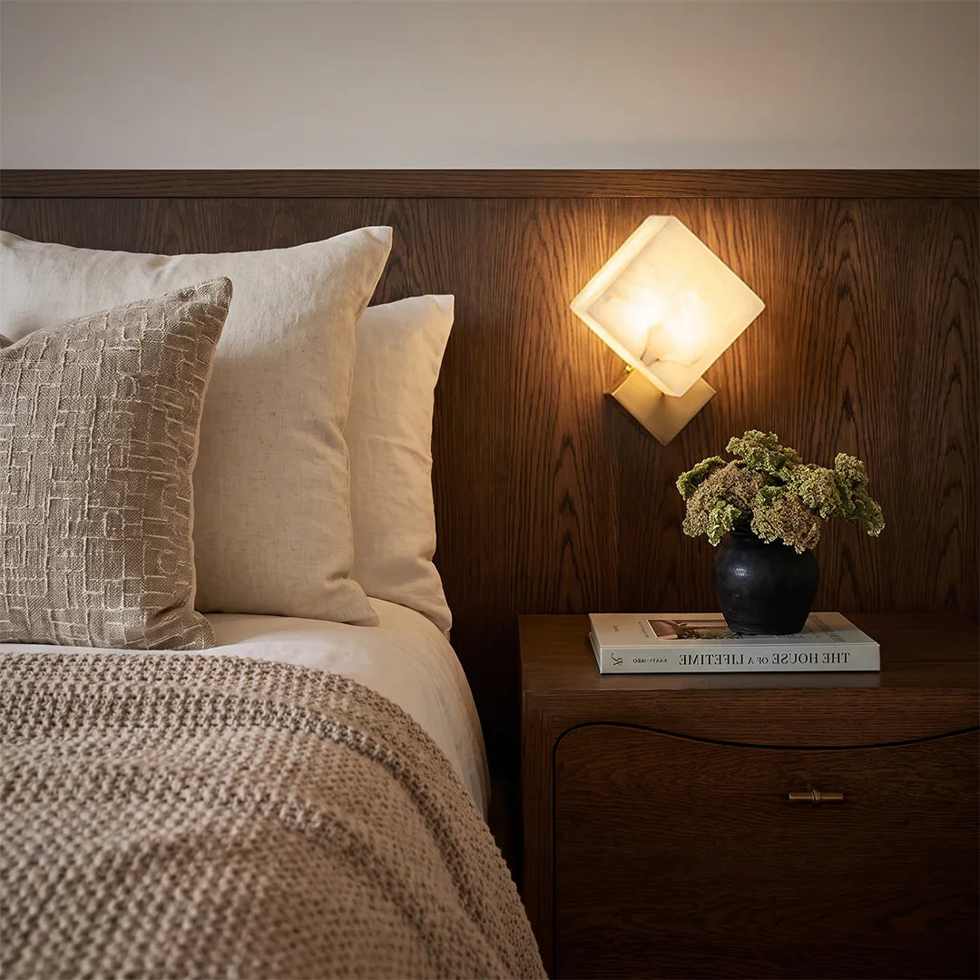 Geometric wall sconce mounted above a nightstand beside a bed in a warm wood-accent bedroom.