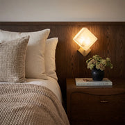 Geometric wall sconce mounted above a nightstand beside a bed in a warm wood-accent bedroom.