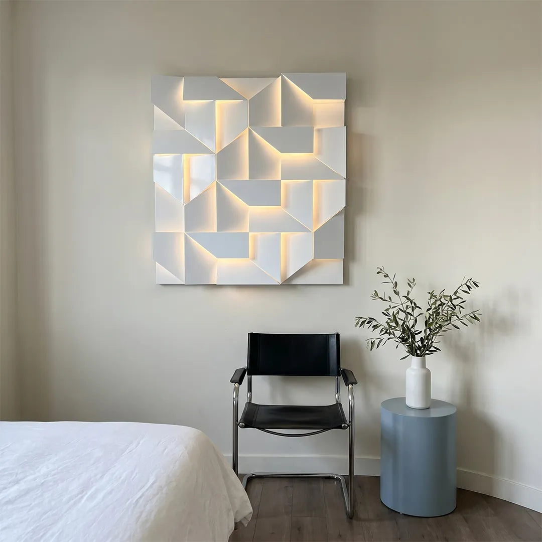 Geometric wall sconce light with sculptural folded panels mounted above a chair in a minimalist bedroom corner.