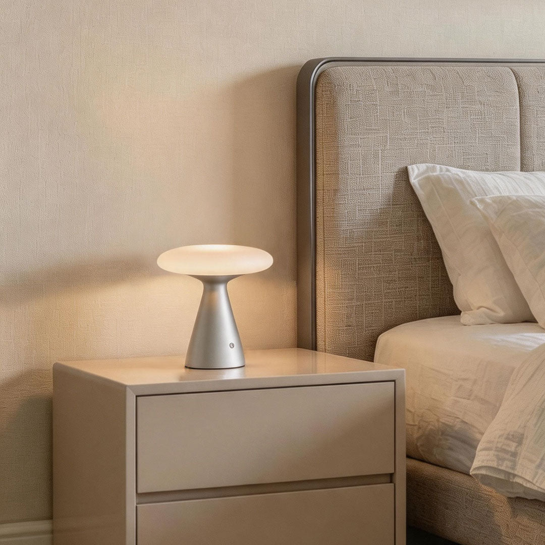 Glass mushroom table lamp displayed on a bedside table next to a soft upholstered bed in a neutral bedroom.