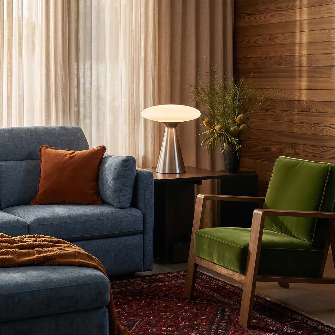 Glass mushroom table lamp placed beside a blue sofa and green accent chair in a warm modern living room.