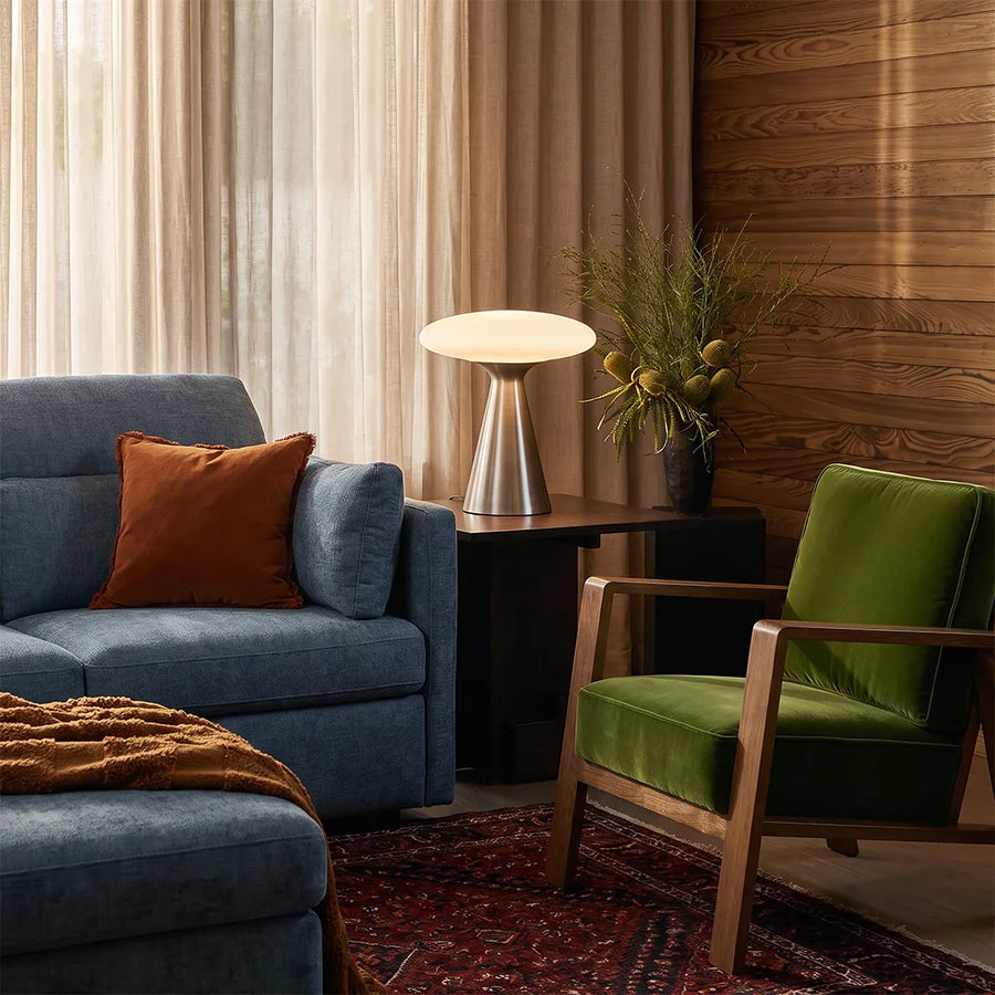 Glass mushroom table lamp placed beside a blue sofa and green accent chair in a warm modern living room.