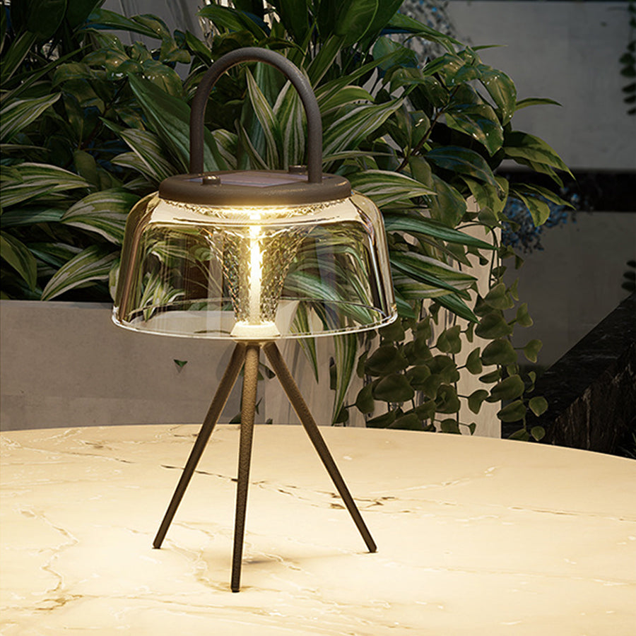 Single glass table lamp with tripod base positioned on surface with soft ambient lighting and plants