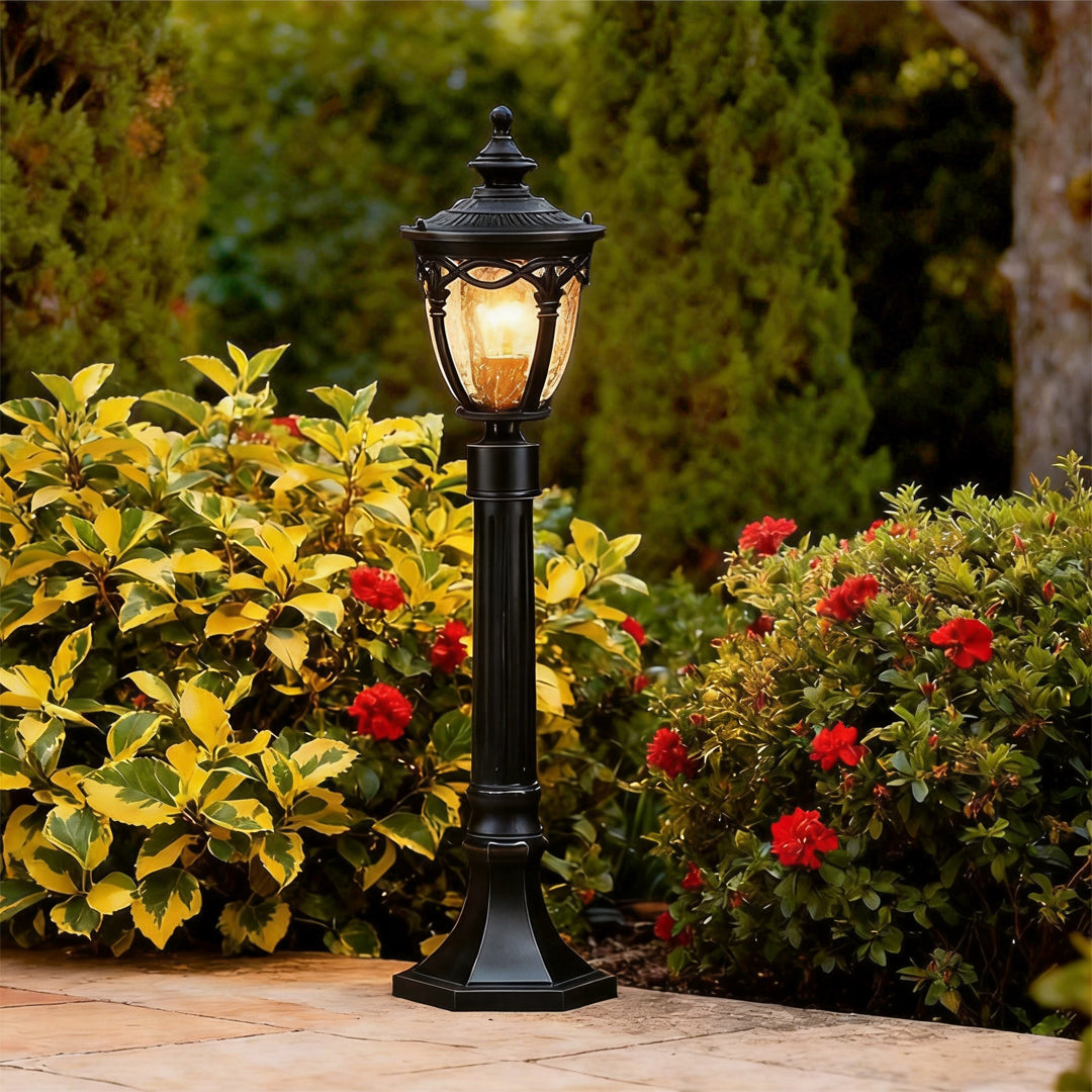 Glowing black outdoor light post illuminating a garden path near vibrant yellow and red plants.