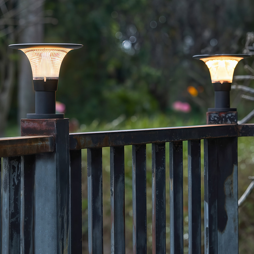 Warmly glowing fence post cap lights with a 'halo' solar panel, shown mounted on a dark metal fence at dusk.