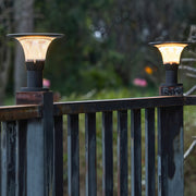 Warmly glowing fence post cap lights with a 'halo' solar panel, shown mounted on a dark metal fence at dusk.