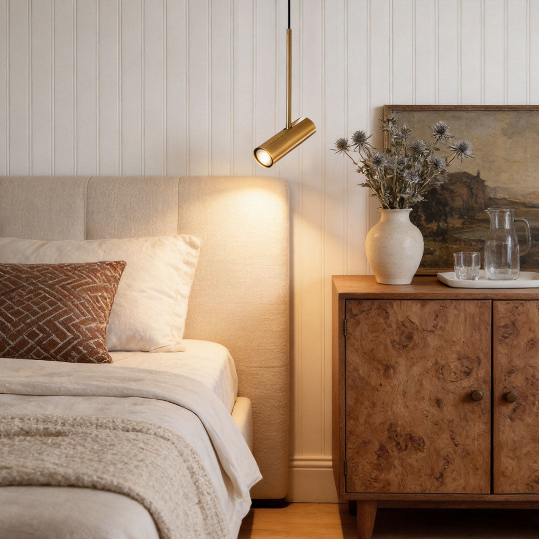 Gold adjustable cylindrical pendant light angled near a bed and a vintage wood cabinets.
