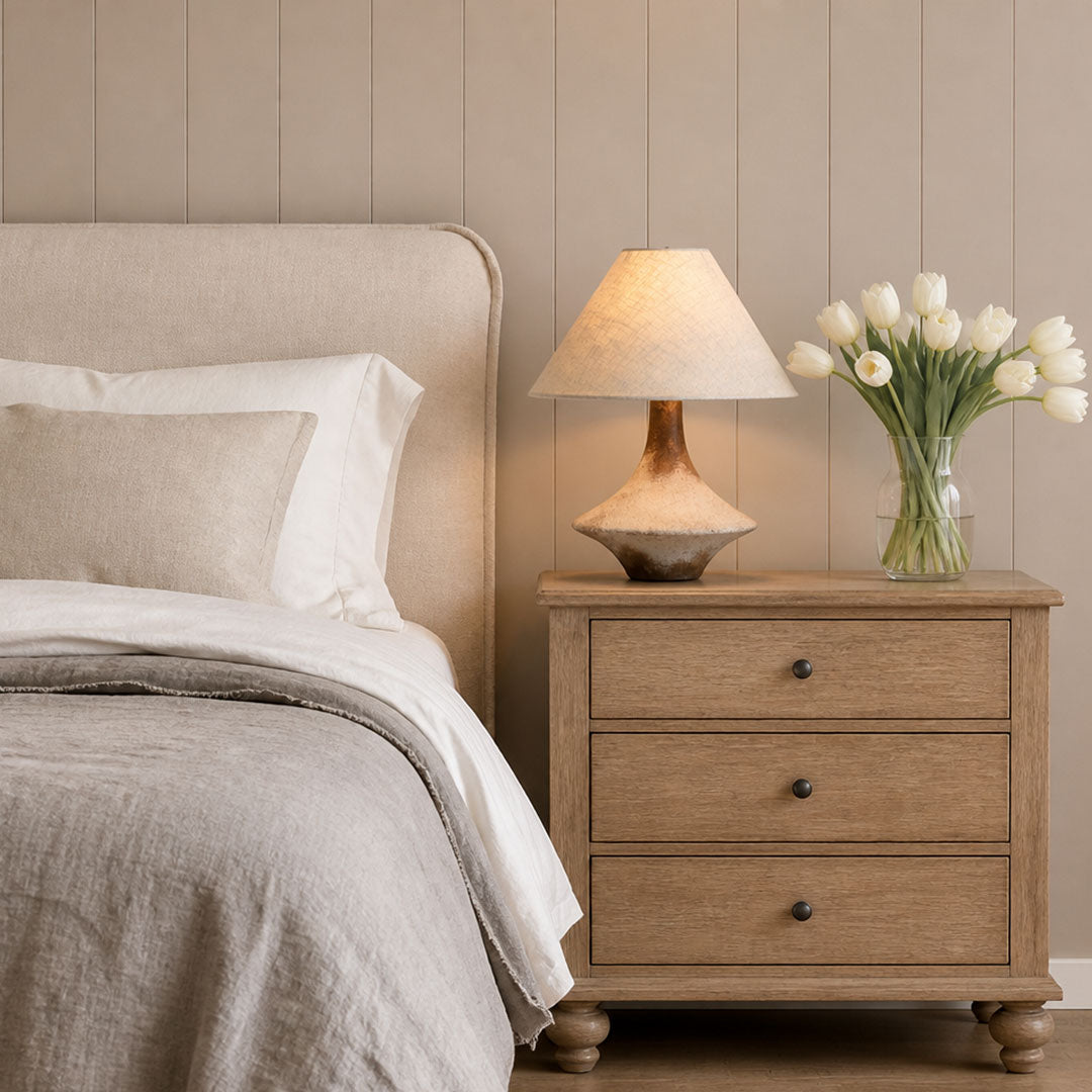 Gray ceramic table lamp on a bedside nightstand beside a neutral upholstered bed in a soft, calming bedroom.