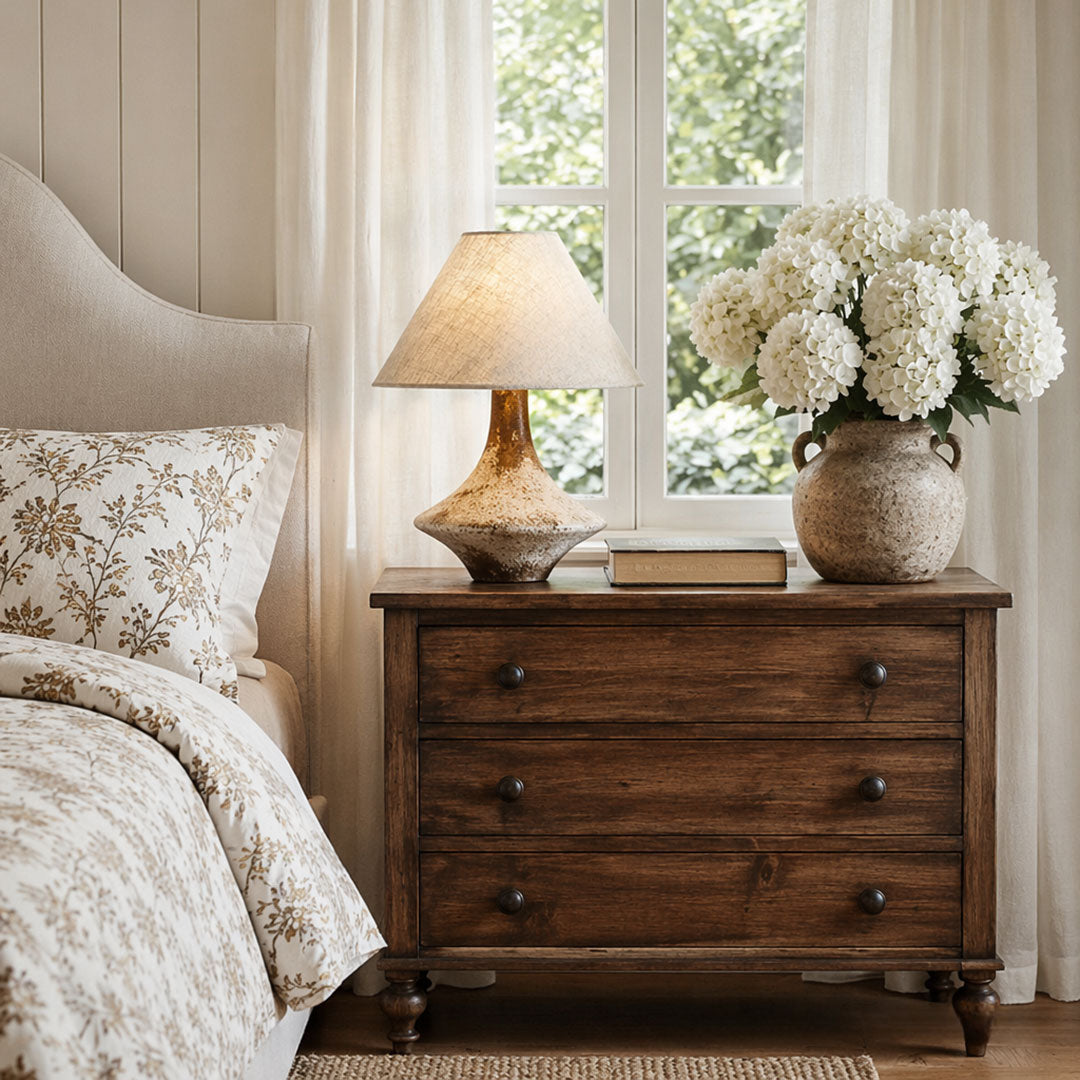 Gray ceramic table lamp styled on a dark wood dresser beside a bed and window with fresh white flowers.