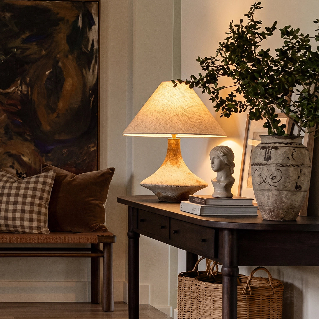 Gray ceramic table lamp on a dark wood side table, styled with books and greenery in a warm living room corner.