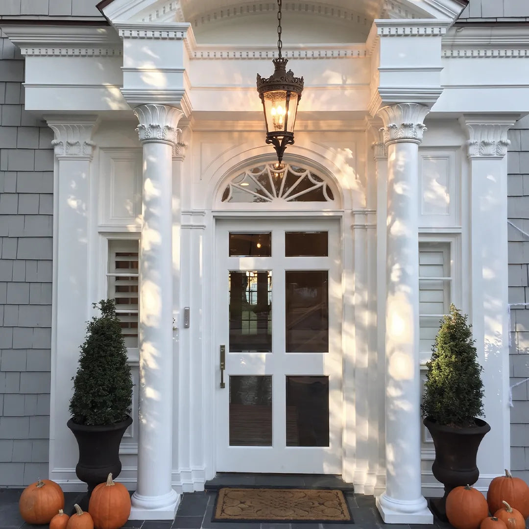 Hanging outdoor porch light above a front entry with white columns, black planters, and pumpkins.