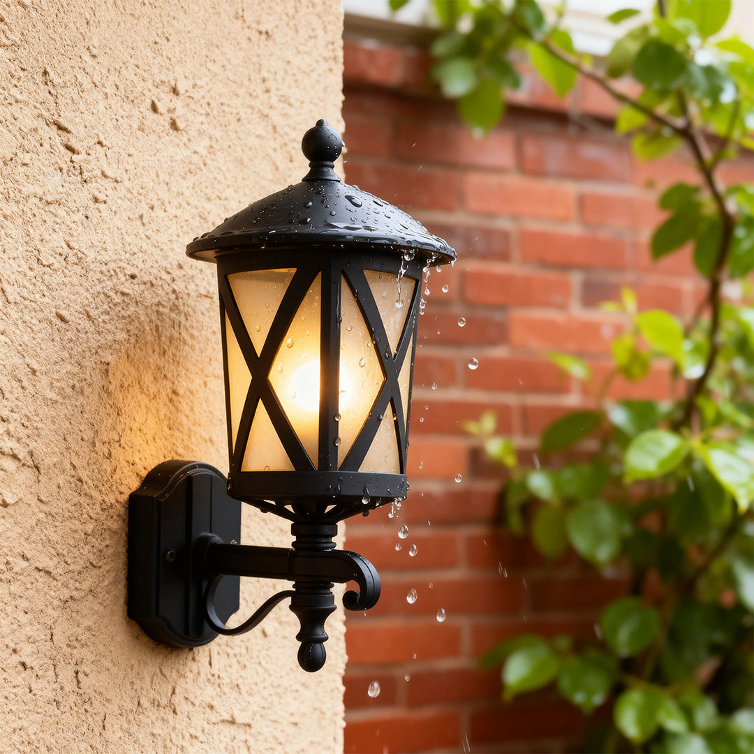 Illuminated outdoor black wall light mounted on textured stucco beside red brick with green foliage