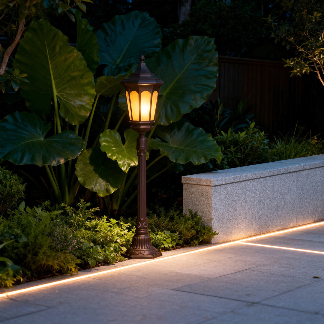 Illuminated post path light casting golden glow along curved concrete walkway at night with tropical plants
