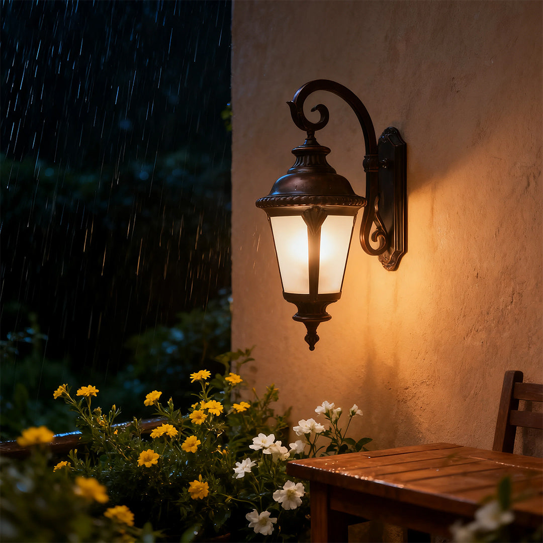 Illuminated vintage outdoor wall sconce glowing warmly on brown exterior beside yellow flowers at dusk