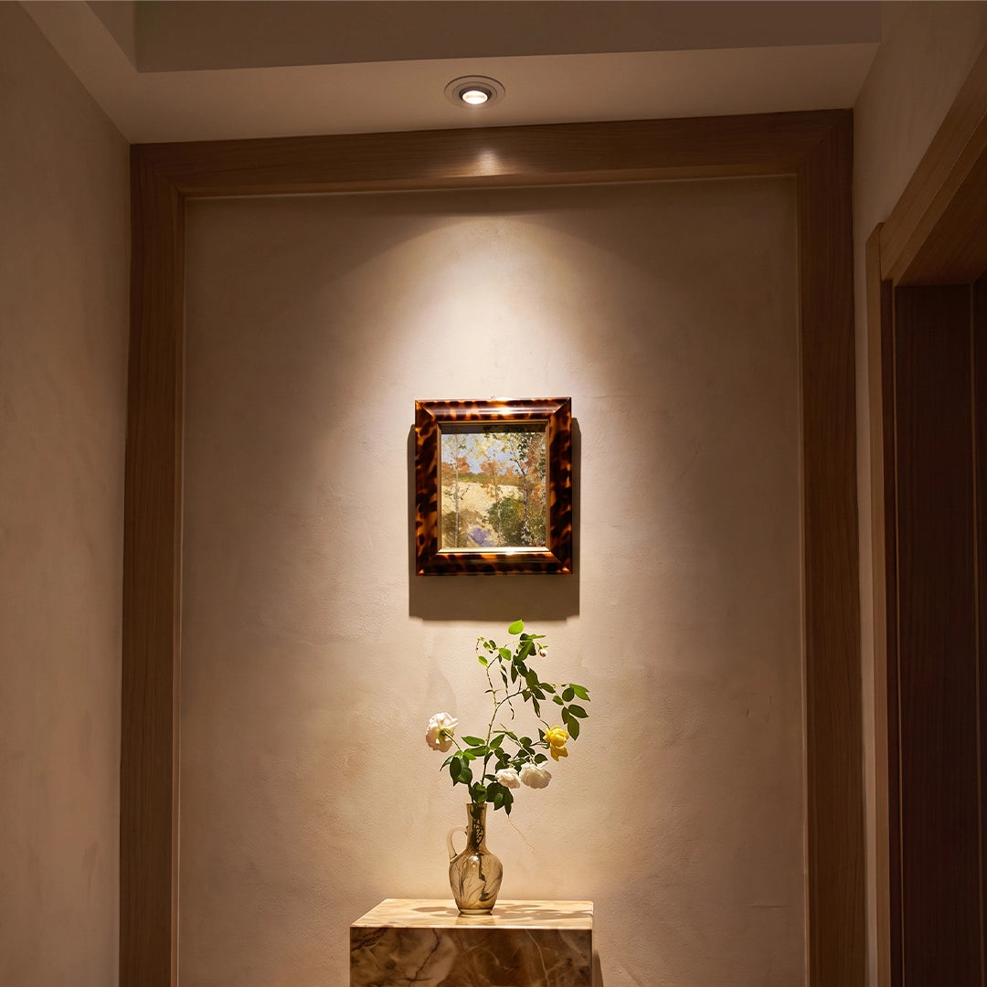 Indoor spotlights illuminating a framed artwork and small table in a warm hallway niche.