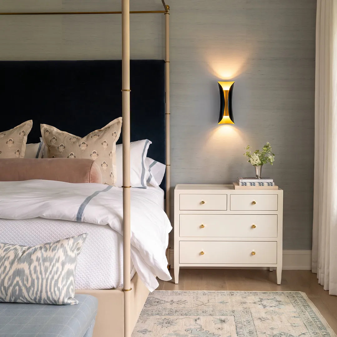 Indoor up down wall lights glowing above a nightstand in a cozy bedroom with a canopy bed.