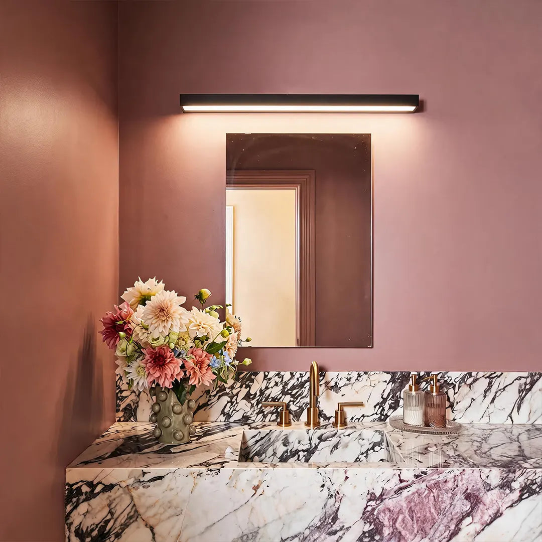 Indoor wall washer light mounted above a bathroom mirror, illuminating a marble vanity against a mauve wall.
