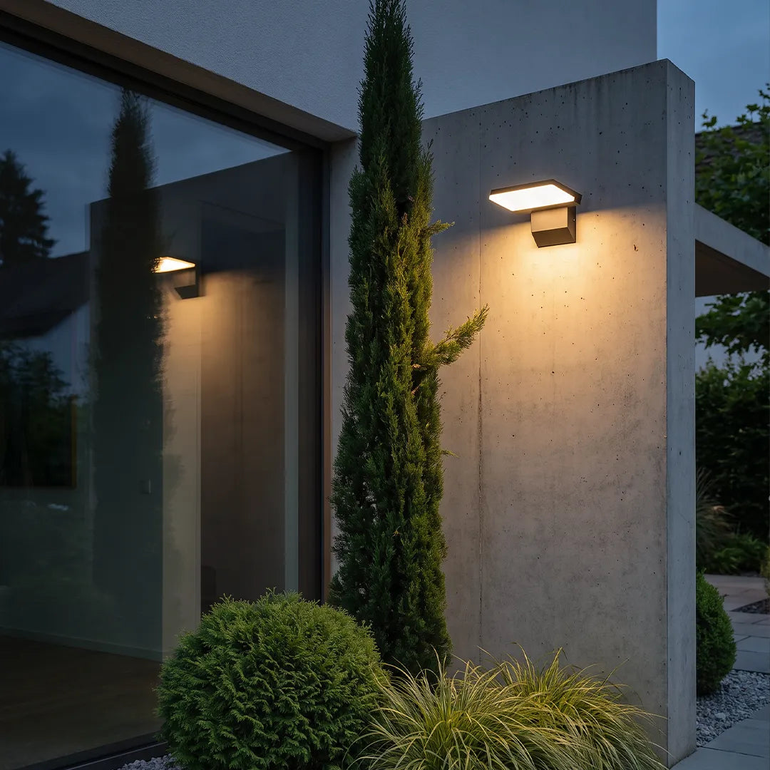 Industrial wall light mounted on a concrete exterior wall, casting warm light beside tall landscaping.
