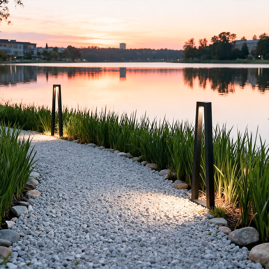 Outdoor lighting for landscaping illuminating a lakeside gravel pathway with modern bollard lights at sunset.