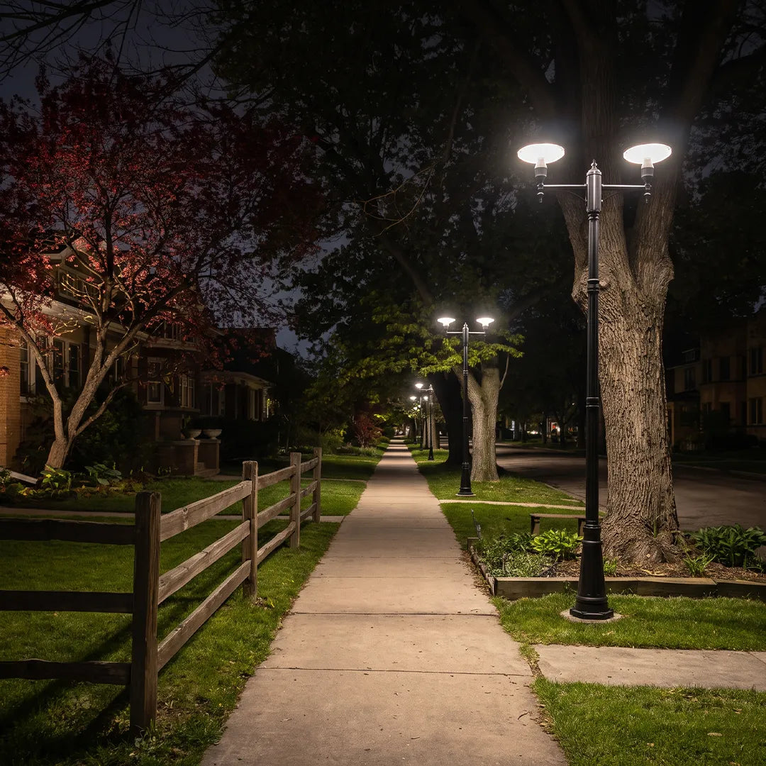 Lamp post lighting illuminating a residential sidewalk lined with trees and fencing at night.