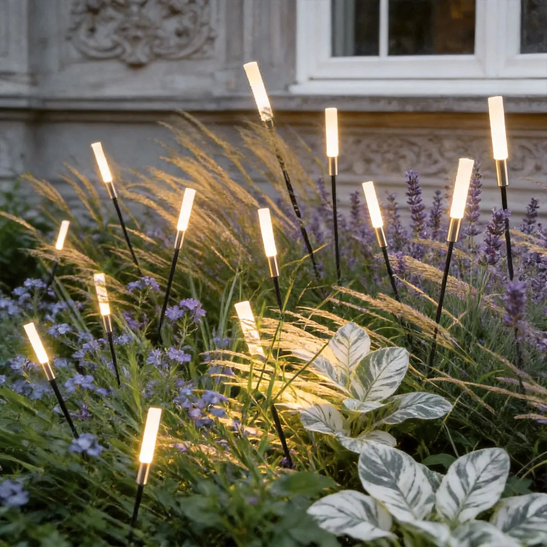 Landscape light spikes arranged among layered plants and grasses beside a home exterior wall.