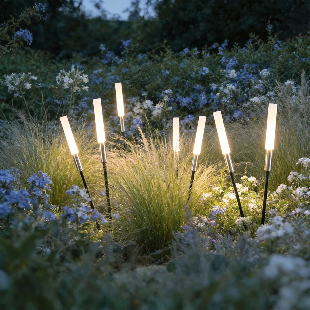 Landscape light spikes glowing among ornamental grasses and blue flowers in a landscaped garden bed.