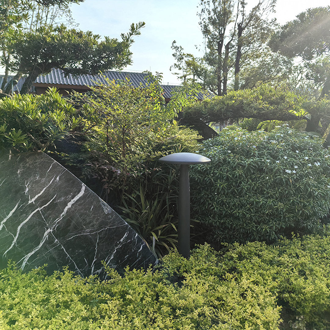 Black mushroom bollard light installed along a landscaped garden with green shrubs and stone walls.