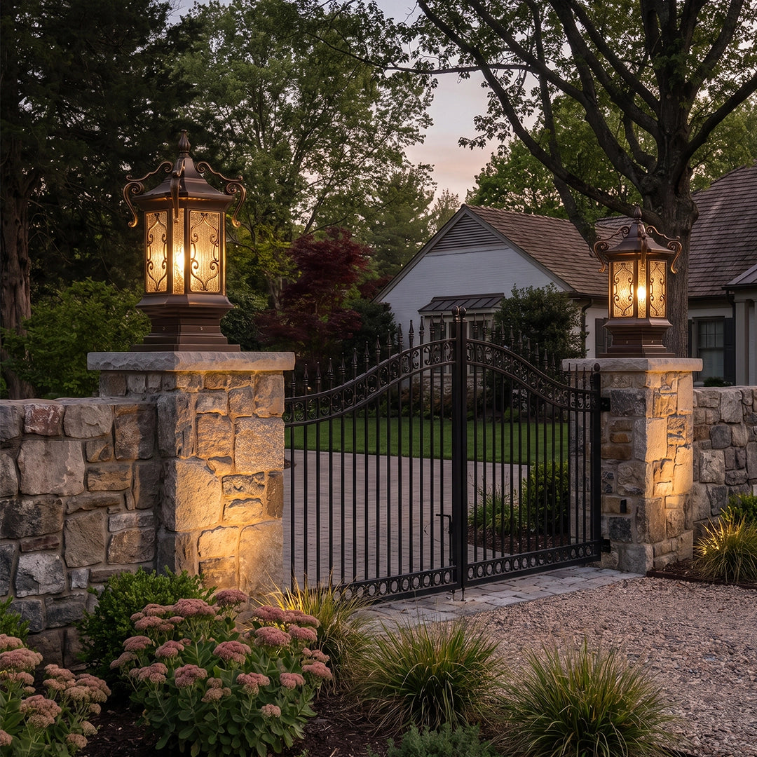 Lantern pillar lights installed on stone gate columns framing a black iron driveway gate at dusk.