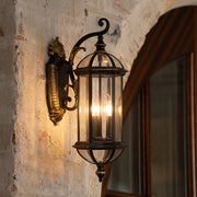Lantern style outdoor wall light in a vintage black and gold finish mounted on a textured exterior wall, shown in a close-up porch setting.