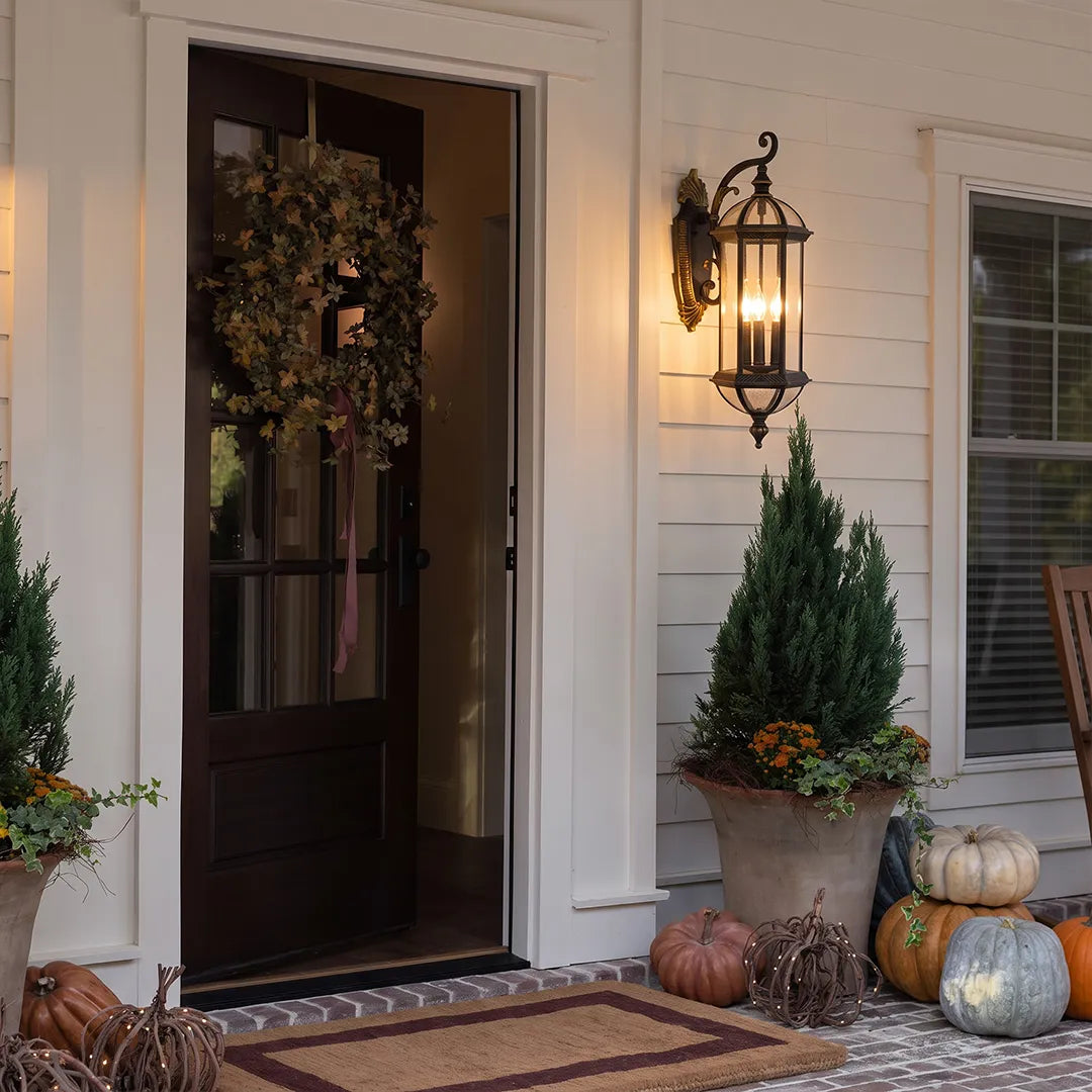 Lantern style outdoor wall light installed beside a dark front door with seasonal decor, adding warm illumination to the porch.