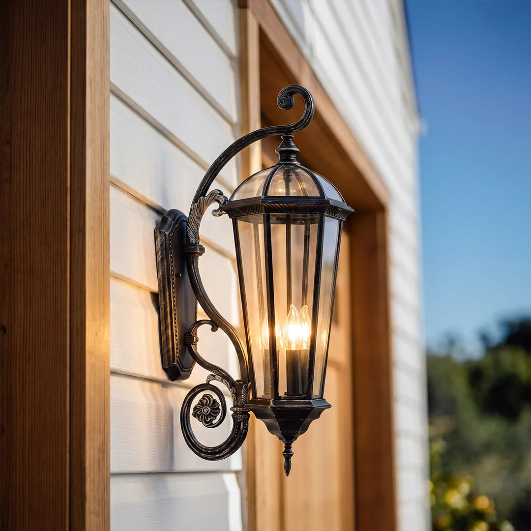 Lantern style wall sconce shown in a close-up exterior view, featuring a scroll arm, clear glass panels, and warm candle-style bulbs.