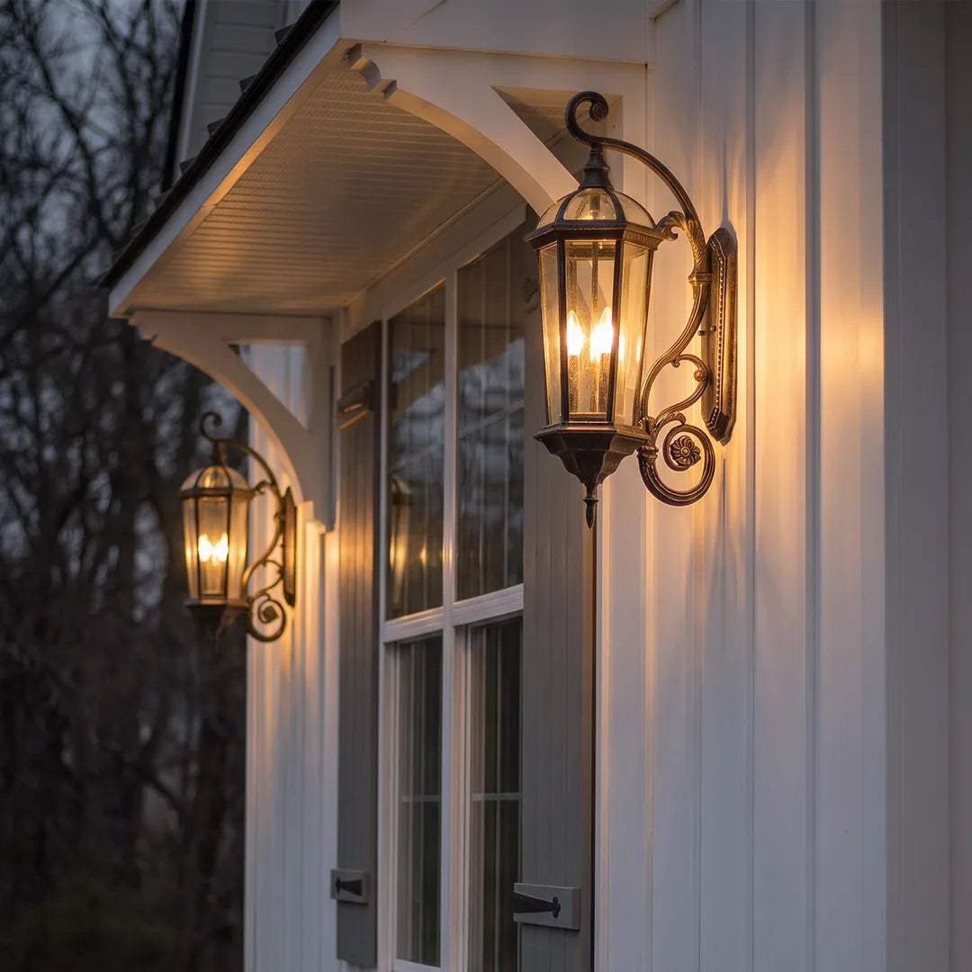 Lantern style wall sconce mounted on a white exterior wall under a porch roof, casting a warm glow beside the entryway.