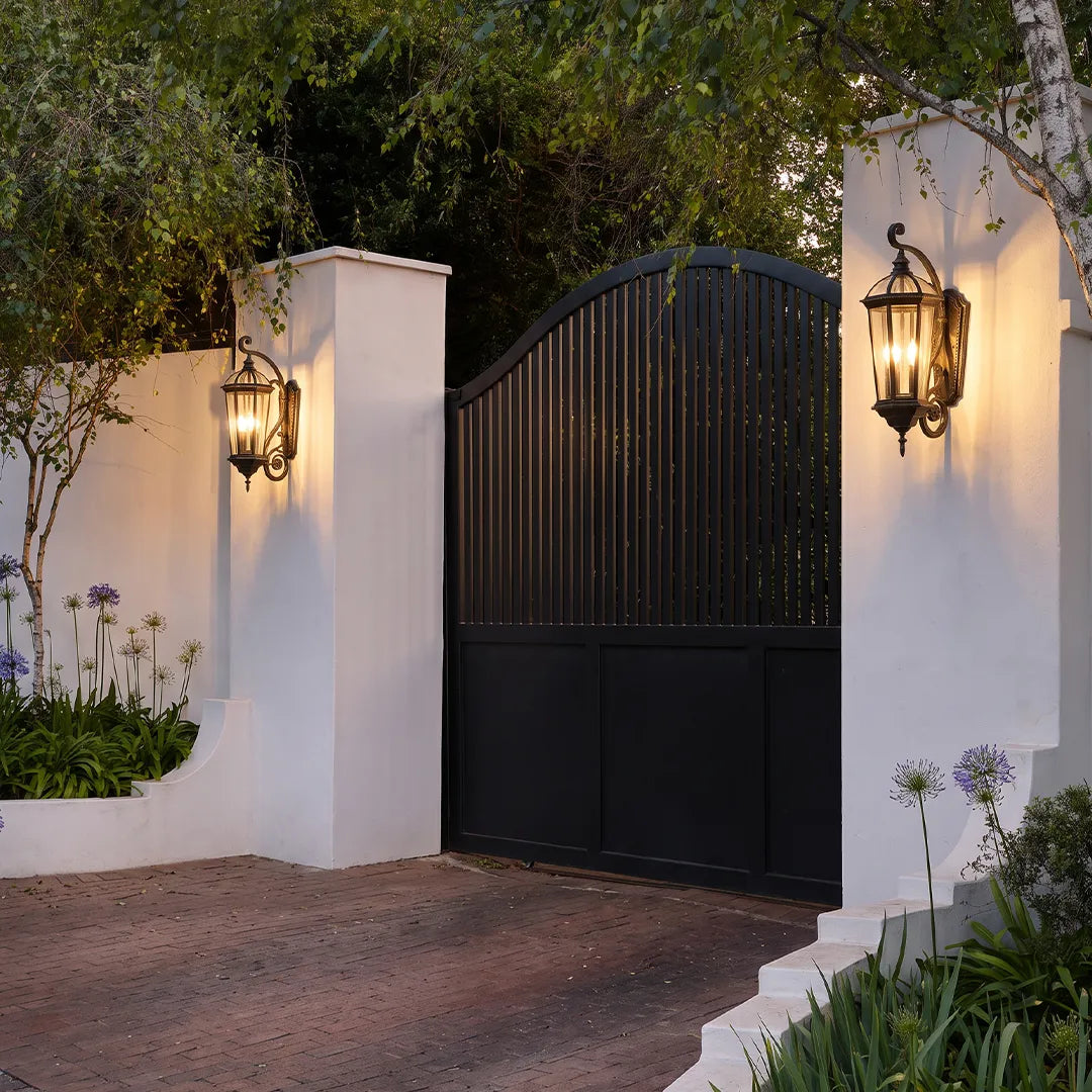 Lantern style wall sconces mounted on gate pillars, illuminating a black entry gate with classic outdoor charm.