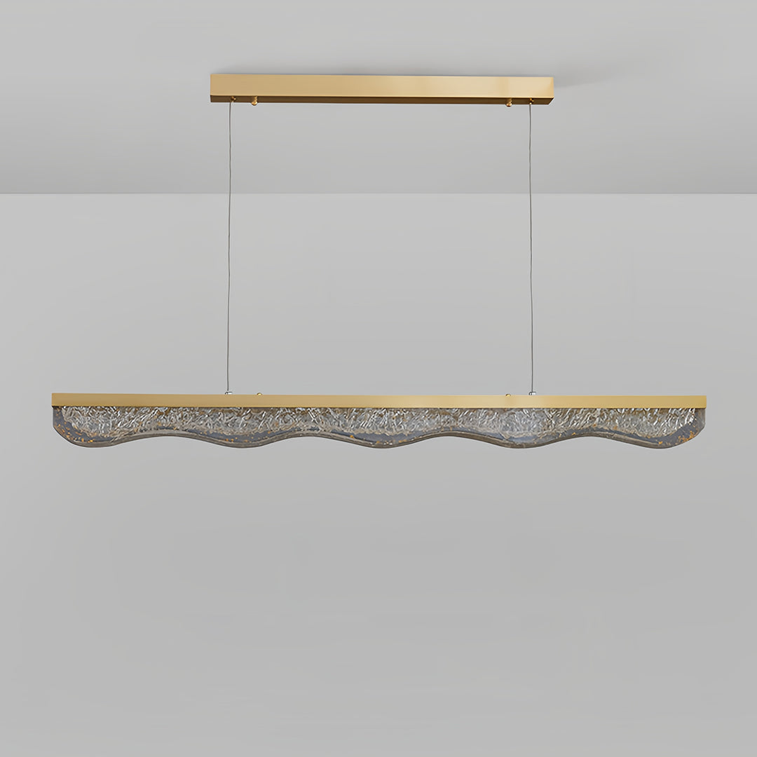 Large gold pendant light showing linear form with textured crystal element on neutral backdrop.