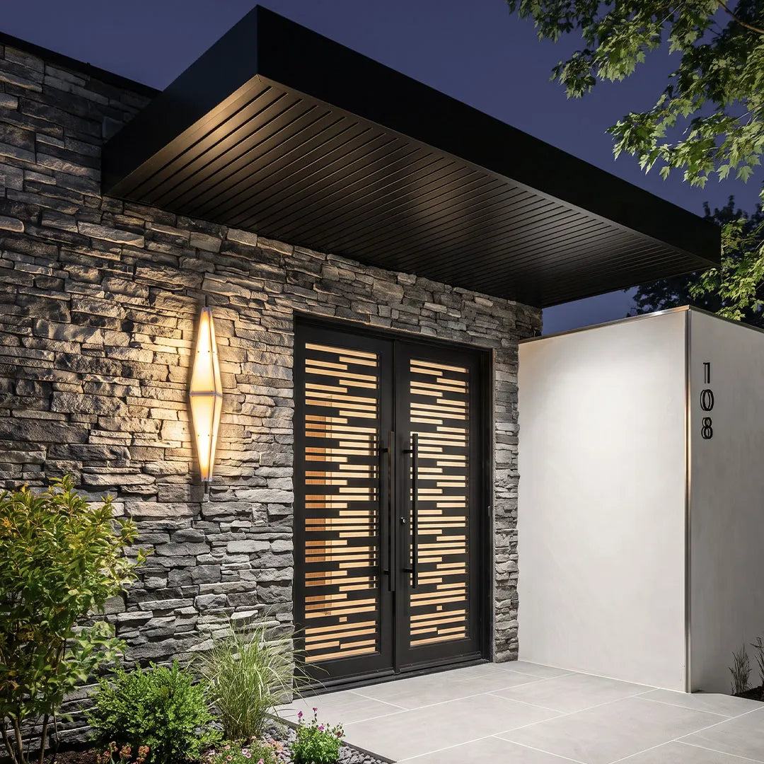Large outdoor sconces illuminating a modern stone exterior beside a black slatted gate at night.