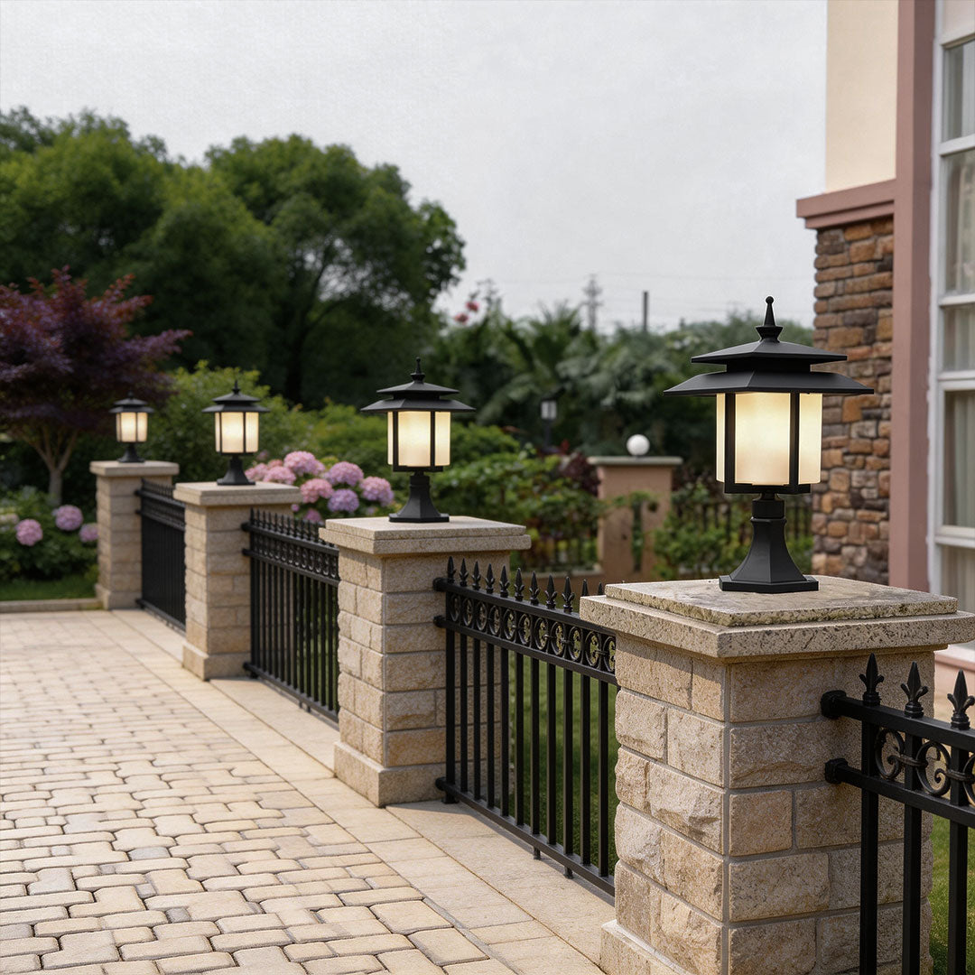 Modern large pillar light outdoor installed along a brick pathway with decorative plants and black fence.