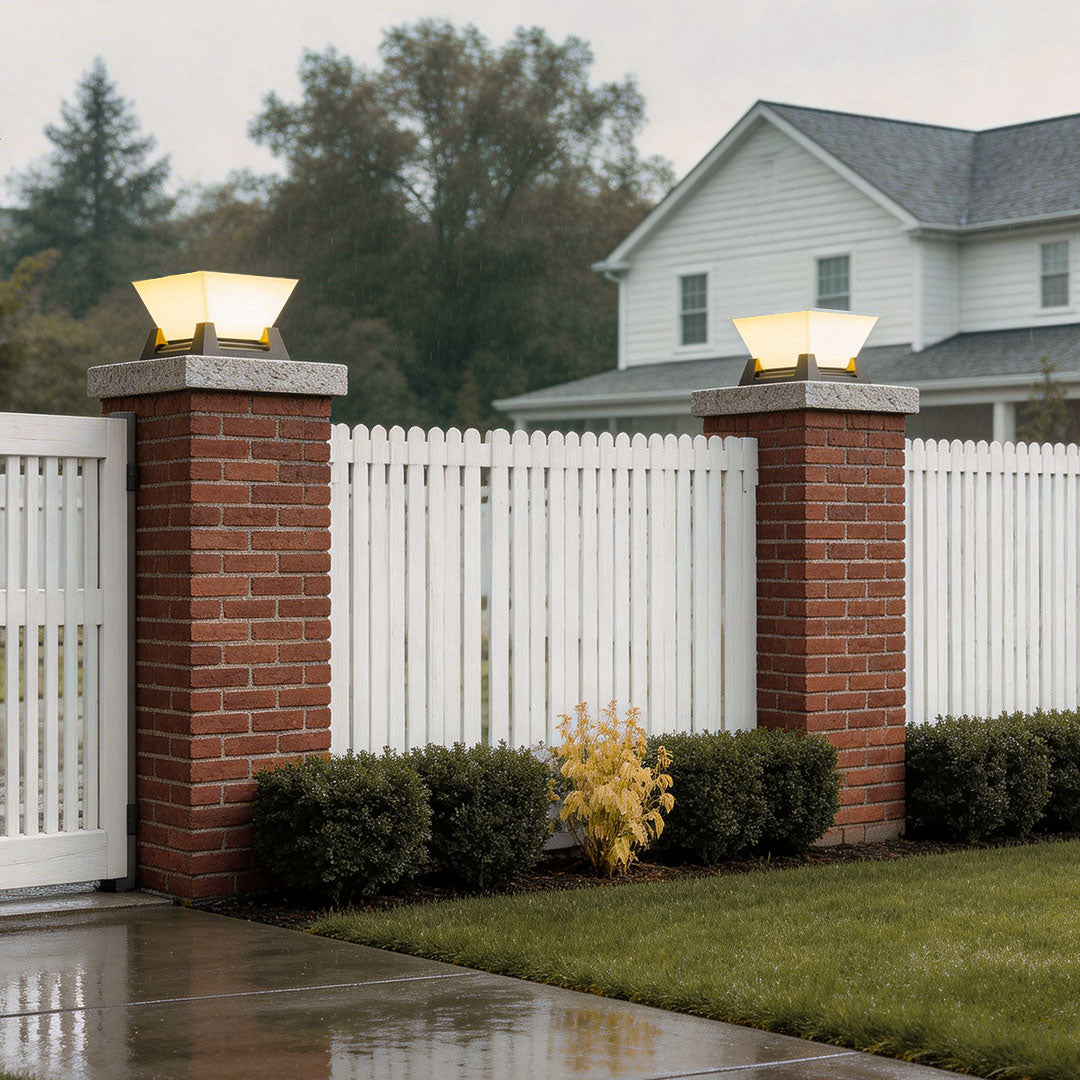 Large pillar lights installed on brick pillars beside a white picket fence and lush garden.