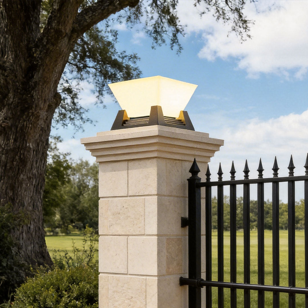 Elegant large pillar lights on a stone pillar beside a black metal fence and mature tree.