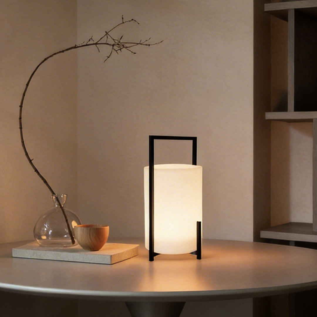 LED cylinder light fixture with a sleek black frame, placed beside decorative plants.