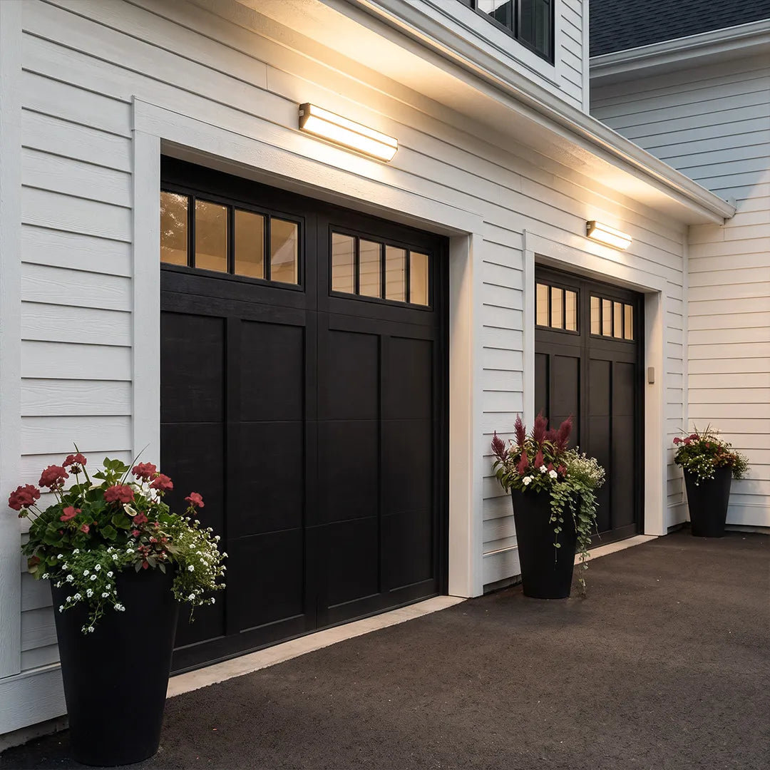 LED linear wall sconces installed above black garage doors on a white house exterior.