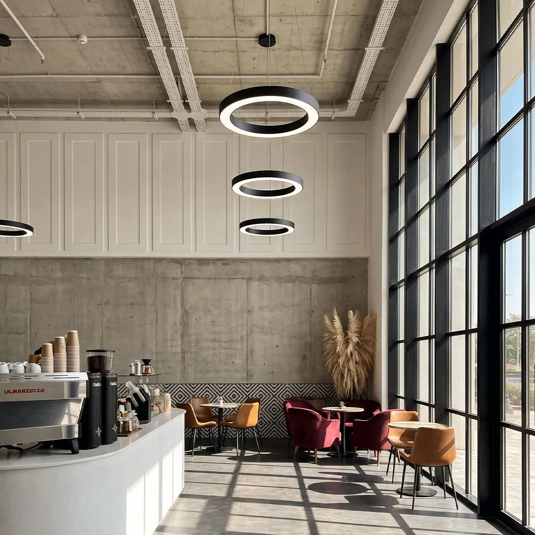 LED pendant ring ceiling light hanging above a contemporary office lounge with tall windows and a coffee bar.