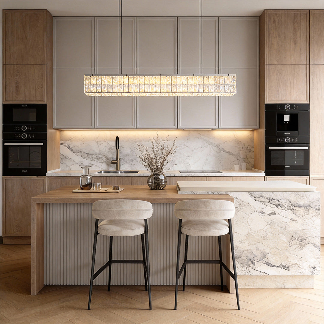 Linear crystal light fixture illuminating a modern kitchen island with marble countertops and wooden bar stools.